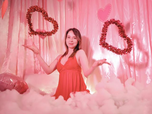 AmbeerWood, uma mulher atraente com longos cabelos escuros, veste um vestido vermelho, estando em pé em neve fofa com tons de rosa, segurando algo vermelho. coroas em formato de coração nas duas mãos, com um coração rosa atrás dela.