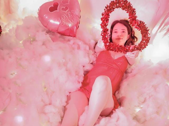 AmbeerWood, lying in a pink-tinted, fluffy white bed, wearing a red latex dress and white stockings. Red tinsel wreath on head, shiny pink heart with "LOVE YOU" text nearby. Fucking sexy.