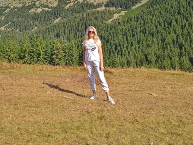 GladisBelle está de pie en un prado cubierto de hierba, vestida con una camiseta blanca sin mangas y pantalones, y con zapatillas blancas. Está mirando hacia adelante con una ligera sonrisa, rodeada de colinas boscosas y densas. Soleado, informal..