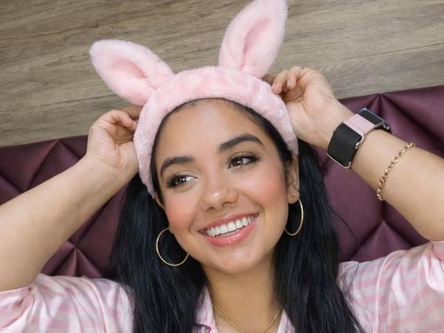 DanMartines, con piel morena, cabello negro y ojos marrones, sonriendo ampliamente, lleva orejas de conejo rosas, aros de oro y un atuendo rosa. Camisa de satén, y reloj inteligente negro, manos ajustando las orejas, ambiente sensual..