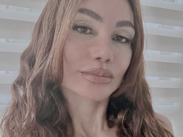 Gros plan de Paulatina, une femme aux longs cheveux bruns, aux lèvres pulpeuses et aux sourcils épais, qui regarde directement l'appareil photo. Un sourire discret. Un éclairage doux met en valeur son visage. Une tension sexuelle est palpable..