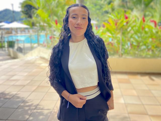 Samantha Roses se tient sous une lumière vive, vêtue d'un débardeur blanc et d'un blazer noir, les mains devant elle. Elle a les cheveux noirs, bouclés et humides, et un sourire discret. L'arrière-plan comprend une verdure floue et des fleurs rouges..