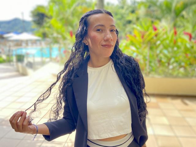 SamanthaRoses with long, wavy black hair, wearing a black blazer over a white crop top, standing outdoors, smiling slightly, green foliage and sunlight in the background, casual, confident pose.