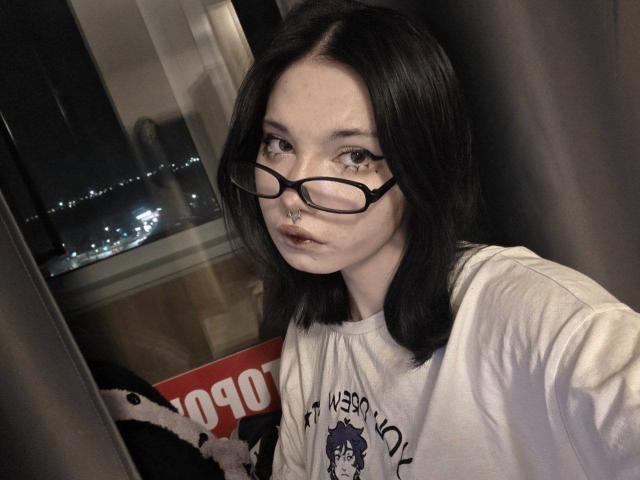 ShayHedglin, con la pelle chiara e i capelli neri, indossa occhiali e una maglietta bianca con una grafica, e guarda direttamente la fotocamera. con la bocca leggermente aperta. Tensione sessuale appena percettibile, sfondo scuro..