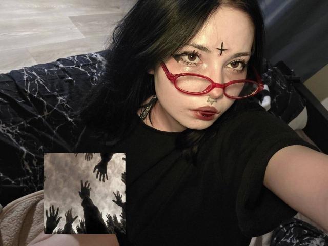 ShayHedglin, una donna dalla pelle chiara, con capelli neri, occhiali rossi, una maglietta nera e una croce nera sulla fronte, che sta guardando. Direttamente verso la telecamera, con un'espressione seducente, tenendo in mano una stampa in bianco e nero con silhouette di alberi..