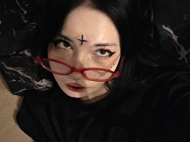 ShayHedglin, met bleke huid en zwart haar, draagt rode brillen en heeft een zwarte ster-tatoeage op haar voorhoofd. Ze kijkt omhoog, haar lippen zijn licht geopend, en je ziet een neusring en donkere lipstick. Extreem intens..
