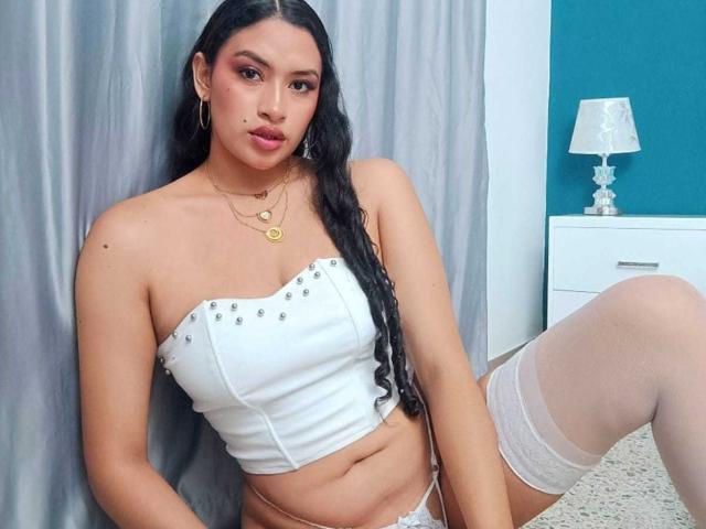 Adalina Vera mit langen, schwarzen Haaren, die ein weißes, trägerloses Oberteil und weiße, spitzenbesetzte Strumpfhosen trägt, lehnt sich an eine grauer Vorhang.
sie schaut direkt in die Kamera und hat ein leichtes Lächeln im Gesicht. Ihr Bein ist angehoben, wodurch ein kleiner Teil ihrer weißen Unterhose zu sehen ist. Ein weißer Nachttisch mit einer Lampe befindet sich im Hintergrund..