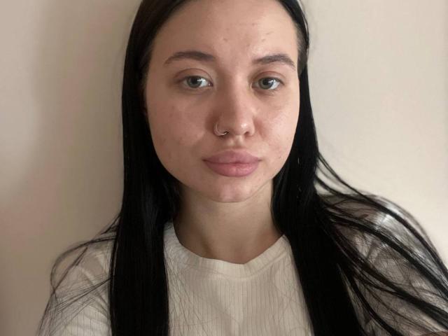 Close-up de AlexaStrei, uma jovem com cabelos longos e pretos, pele clara e um piercing no nariz. Ela está vestindo uma camiseta branca texturizada, olhando diretamente para a câmera com uma expressão neutra. Seus lábios estão levemente franzidos..