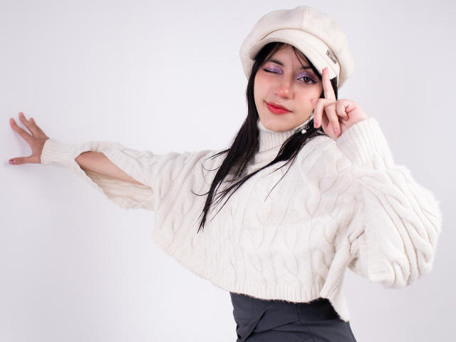 Dafne Rivas, avec ses longs cheveux noirs, porte un pull blanc duveteux et une casquette assortie, un pantalon noir et du rouge à lèvres. Elle se penche en avant, une main touchant son visage, créant une tension sexuelle. Éclairage lumineux.