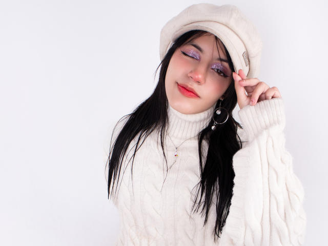 DafneRivas, with long black hair, wears a white knit hat and matching sweater. She touches her hat with one hand, her eyes heavily lined with purple eyeshadow, red lips slightly parted.