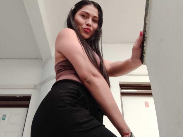 Photograph of ValentinaMoreau, a curvy woman with long black hair, wearing a brown crop top and black pants, leaning against a white wall, looking down with a seductive gaze. Fucking sexy.