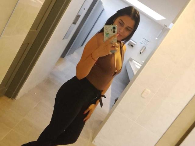 ValentinaMoreau, incroyablement sexy, prend un selfie devant un miroir dans une salle de bain. Elle porte un haut marron, une jupe noire nouée à la taille, et tient un téléphone avec une coque verte. Expression coquine.