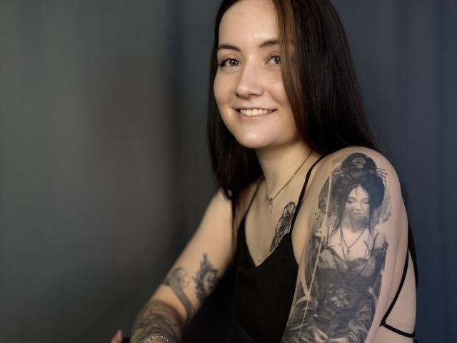 GeraldineHal, avec ses cheveux noirs mi-longs, souriante, portant un débardeur noir, et arborant un tatouage détaillé représentant une femme. avec des poils foncés sur le haut de son bras. Le tatouage est en noir et blanc..