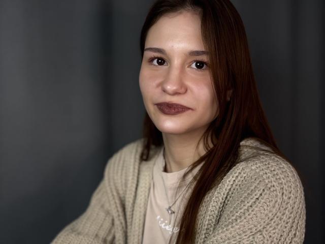 Photograph of a young woman with long brown hair, wearing a beige knit cardigan over a white shirt. She has dark lipstick, a neutral expression, and a subtle glow on her face.