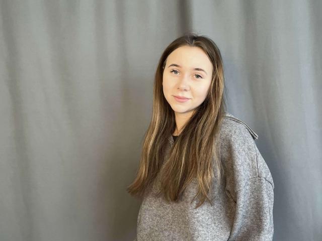 PokBingman, een blanke vrouw met lang bruin haar, die een grijze trui draagt, staat voor een vage, grijze achtergrond. Ze heeft een subtiele, zelfverzekerde glimlach en een ontspannen, aantrekkelijke houding..