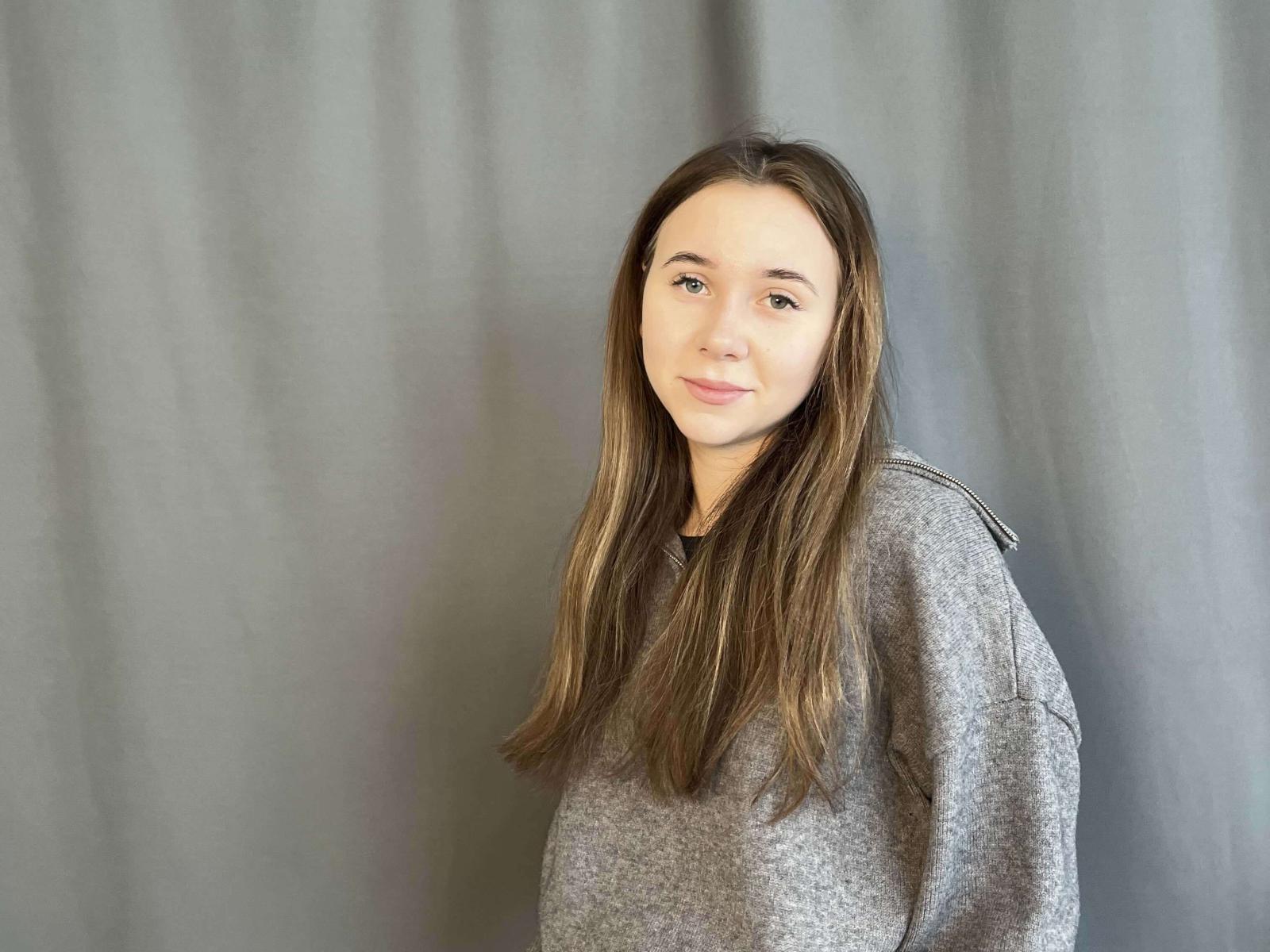 PokBingman, une femme blanche aux longs cheveux bruns, portant un pull gris, se tient devant un fond gris flou. Elle a un sourire subtil et confiant, et une posture détendue et séduisante..