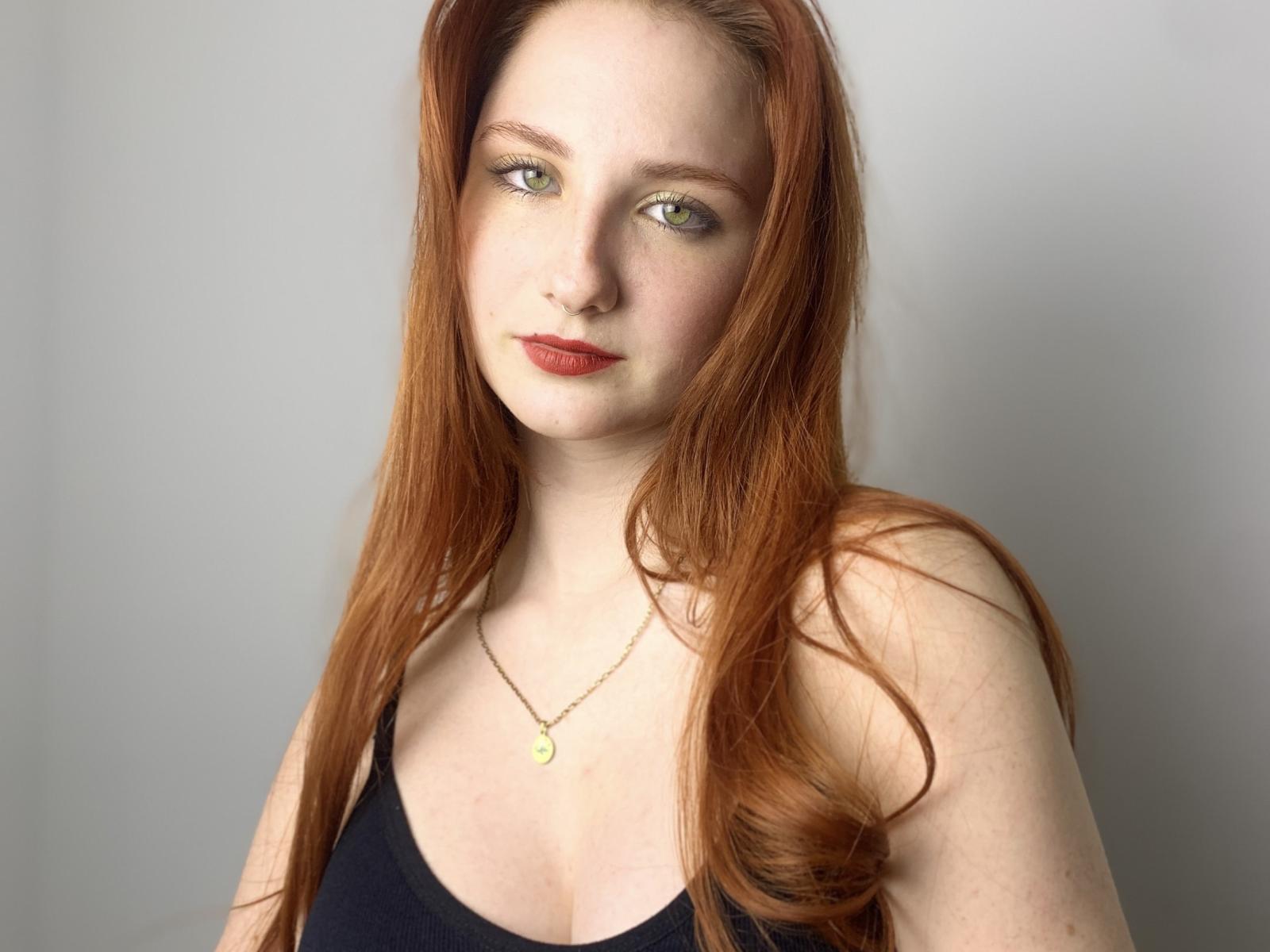 Jennifer, con su largo cabello rojo, piel pálida y ojos verdes, lleva puesto un top negro y mira directamente a la cámara. La luz resalta su rostro, creando una mirada ligeramente sensual e intensa..