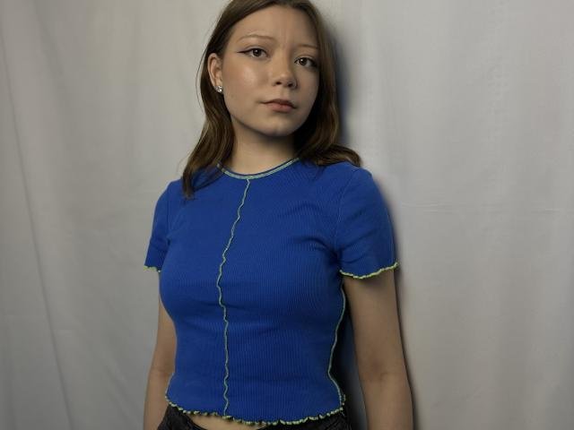 Precious Kinatyan, una giovane donna con la pelle color nocciola e i capelli castani lisci, indossa una maglietta aderente, blu e a coste. bordi gialli, in contrasto con uno sfondo bianco, sottile tensione sessuale, posa informale.