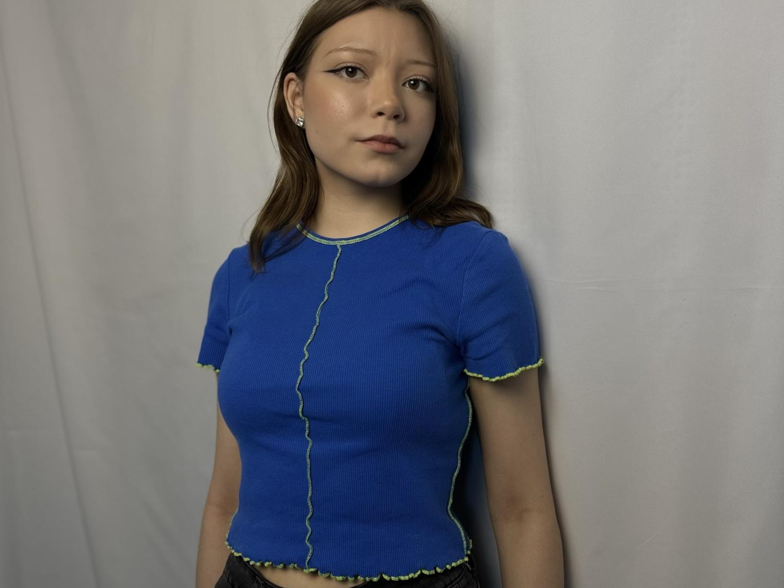 Precious Kinatyan, eine junge Frau mit hellbrauner Haut und glattem, braunem Haar, trägt ein eng anliegendes, blaues, geripptes T-Shirt. gelbe Akzente, vor einem weißen Hintergrund, subtile sexuelle Spannung, lässige Pose.