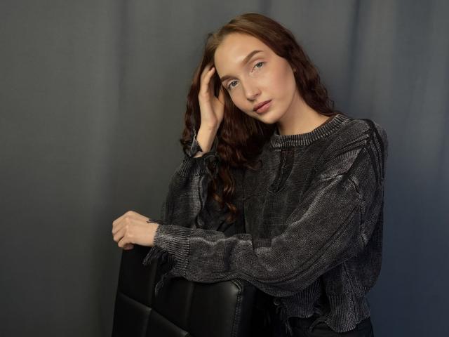 PattyZeidman, with long wavy brown hair, wearing a black textured sweater, leans against a dark backdrop, hand in hair, sultry expression, slight tilt to the head, serious gaze.