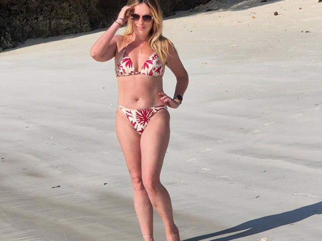 Sofie: Sexo apasionado con un bikini floral rojo y blanco, de pie en una playa iluminada por el sol. Es alta, rubia, con gafas de sol, y está arreglándose el pelo. Hay una sombra en la arena, y la luz del sol es brillante..