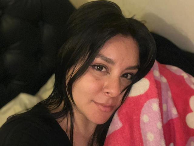Gros plan d'EmaJames, une femme aux longs cheveux noirs, allongée sur un lit, la tête reposant sur une couverture à pois rouges et blancs, regardant la caméra avec un léger sourire. Éclairage doux.
