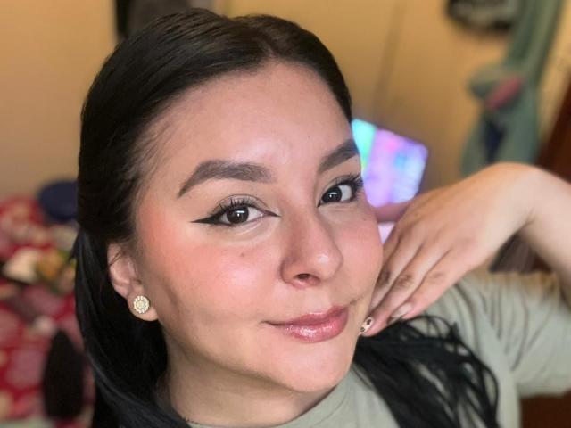 Close-up of EmaJames, a woman with dark hair, wearing a green shirt, touching her cheek with one hand. She has black eyeliner, diamond earrings, and a slight smirk. Background blurred.