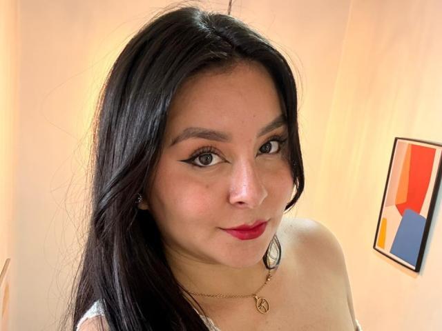 Close-up of EmaJames with long black hair, red lipstick, and black eyeliner. She's wearing a gold necklace and white top. Framed abstract art in the background. Fucking hot, confident smile.