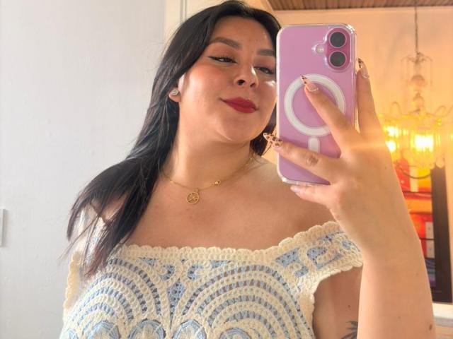 EmaJames, con cabello negro largo, vestida con un vestido azul y blanco de ganchillo con un corte que deja el hombro al descubierto, sosteniendo un teléfono rosa con un anillo. agarre, tomando una selfie con un espejo, sonriendo con lápiz labial rojo.