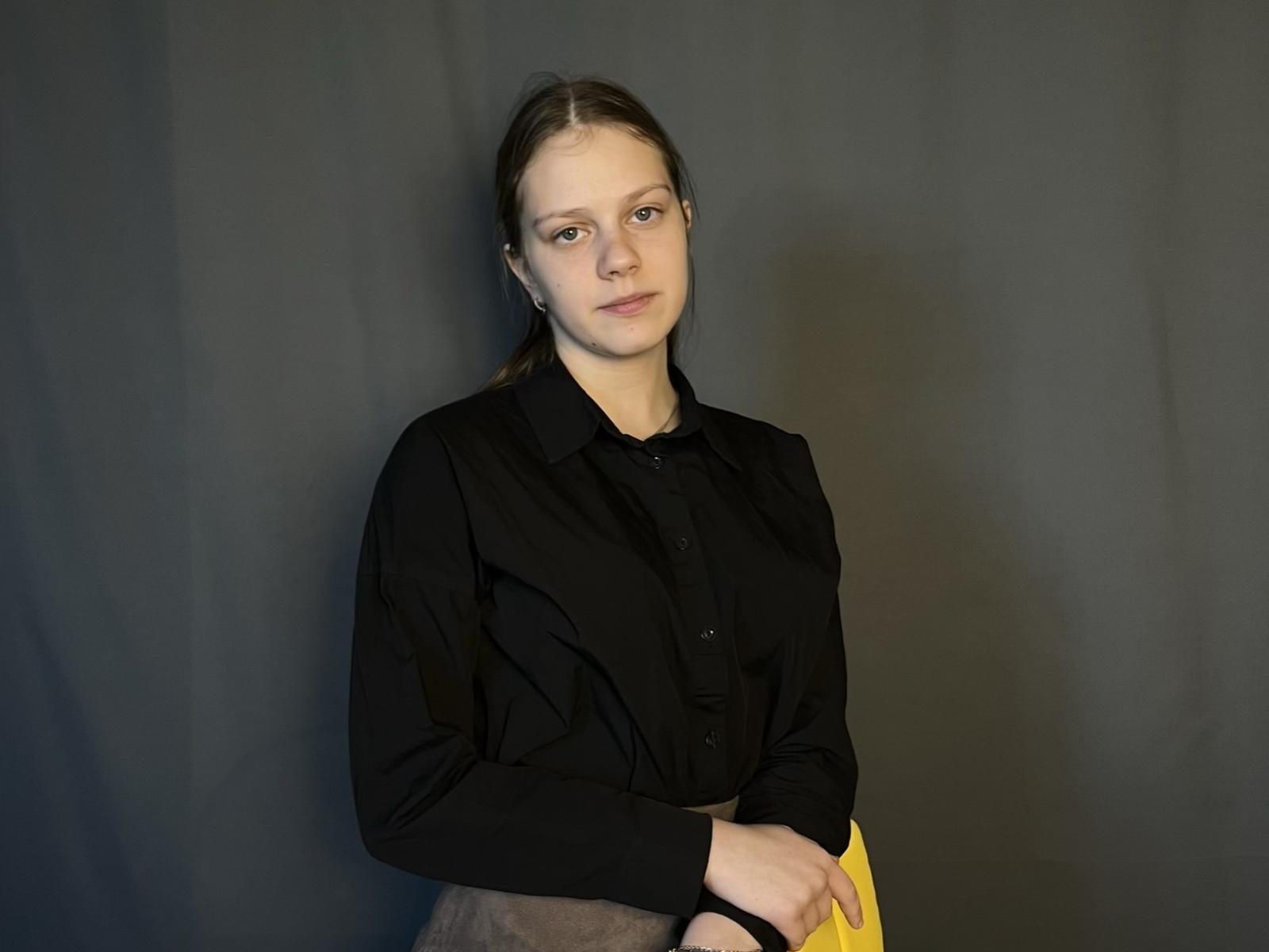 Fotografia de Carlie Hobden, com pele clara, cabelo castanho e uma expressão séria. Ela veste uma camisa preta com botões e segura um tecido amarelo. Fundo preto. Expressão muito séria, sem o pênis visível..