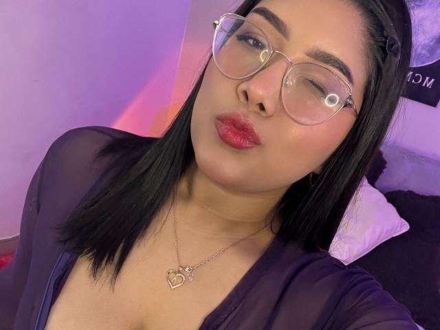 Close-up of AlexaKiim, a sexy Asian woman with black hair, wearing clear glasses and a black top, winking with red lips, heart necklace, purple and pink lighting in the background.