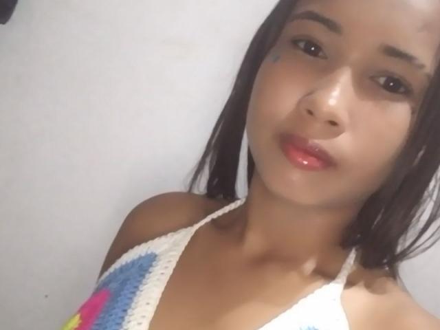 Fotografía de primer plano de SofiaPerezLov, con piel de color marrón medio, cabello negro largo y una pequeña marca azul en su mejilla. Ella lleva un top de halter de ganchillo de colores y tiene los labios pintados de un rojo brillante. Increíblemente atractivo/a..