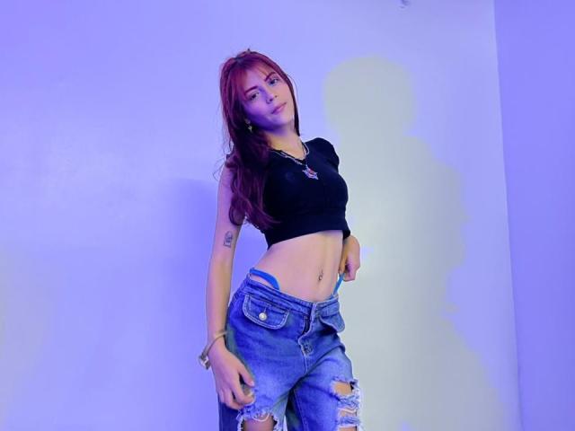 ViioletteRoselle, magra, con la pelle chiara, lunghi capelli rossi, un top corto nero, jeans strappati, in piedi di fronte a uno sfondo bianco illuminato di blu. muro, mani sui fianchi, sicura di sé, sexy, piercing visibile all'ombelico, tatuaggi sulle braccia.