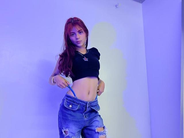 ViioletteRoselle, die an eine weiße Wand gelehnt steht, trägt ein schwarzes Crop-Top und zerrissene blaue Jeans und hält einen blauen Trageriemen. rote Haare, blauer Schimmer, verführerische Pose, selbstbewusster Ausdruck, lässiger Stil.