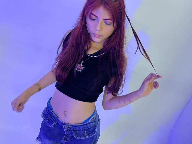 ViioletteRoselle, een slanke jonge vrouw met lang rood haar, draagt een zwarte crop top en blauwe jeans en houdt een haarspeld vast. Haar navel is doorboord, blauwe achtergrond. Verdomd sexy..