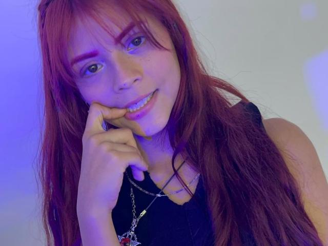 Close-up of ViioletteRoselle, a young woman with long red hair, blue eyes, and a slight smile, biting her finger. She's wearing a black top with a silver necklace. Fucking sexy.