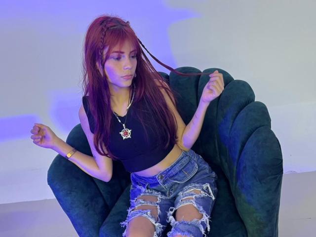 ViioletteRoselle, una donna esile con lunghi capelli rossi, è seduta su una poltrona nera e imbottita, indossando un top corto nero e jeans strappati. pantaloncini di jeans, con i capelli raccolti, illuminata da luci blu. La sua espressione è rilassata.
.