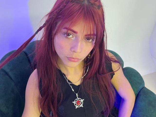 ViioletteRoselle a de longs cheveux roux et indisciplinés, une peau pâle et des yeux marrons. Elle porte un haut noir avec un pendentif rouge et blanc. Elle est assise sur un canapé vert, regardant directement la caméra avec une bouche légèrement ouverte..