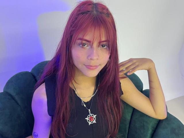 ViioletteRoselle, con lunghi capelli rossi e frangia, è seduta su un divano verde scuro, indossando una canotta nera e un accessorio argentato. collana con un ciondolo rosso.
illuminazione blu sullo sfondo.