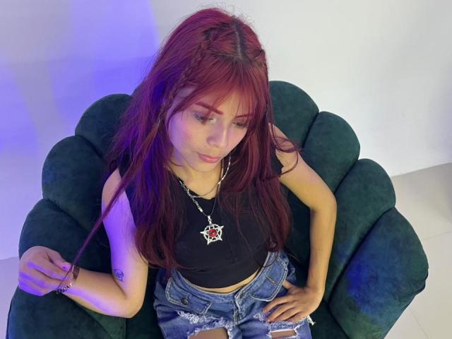 ViioletteRoselle, una donna seducente con lunghi capelli rossi, è seduta su una poltrona nera e imbottita, indossando una maglietta nera e jeans strappati. jeans, con un'espressione sottile e seducente, guardando verso il basso. La luce blu esalta la sua pelle
.