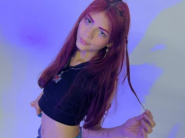 ViioletteRoselle hat langes, rotes Haar, trägt ein schwarzes Crop-Top und hält mit ihrer rechten Hand eine Haarsträhne fest. Blauer Hintergrund. Sie hat ein leichtes Lächeln und schaut direkt in die Kamera..