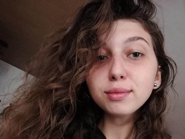 Nahaufnahme von Sanella, einer jungen Frau mit zerzaustem braunen Haar, heller Haut und kleinen Ohrringen. Sie schaut direkt in die Kamera und trägt ein leichtes Lächeln, sie trägt ein schwarzes Oberteil. Total süß..