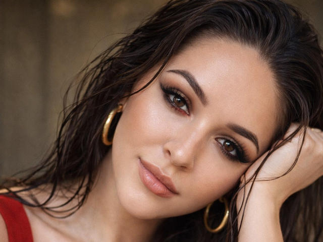 Fotografia ravvicinata di AquaLady con capelli neri bagnati, orecchini a cerchio dorati e trucco occhi smokey. Lei appoggia la testa sulla mano, guardando in modo seducente con un sorriso appena accennato. Molto attraente..