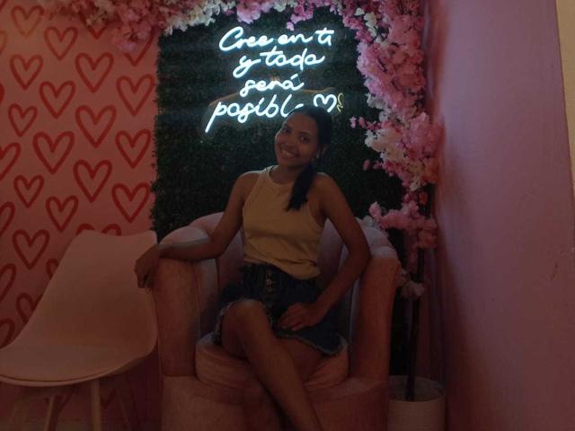 MelaniaVelasquez sits on a pink chair, wearing a yellow top and black shorts, under neon "Queremos que te passei um pól" sign, surrounded by pink flowers and heart-patterned wall. Dim lighting.