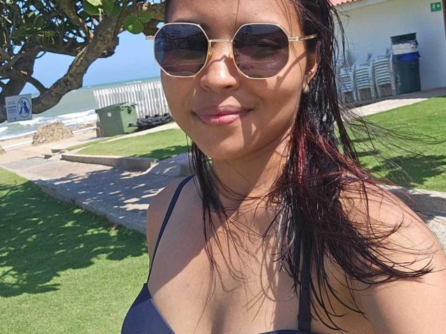 Melania Velasquez, com cabelo escuro, usando óculos de sol e uma blusa preta de alça fina, está em um dia ensolarado ao ar livre. Grama verde, árvore, cadeiras brancas e uma lixeira verde ao fundo. Sorrindo levemente.