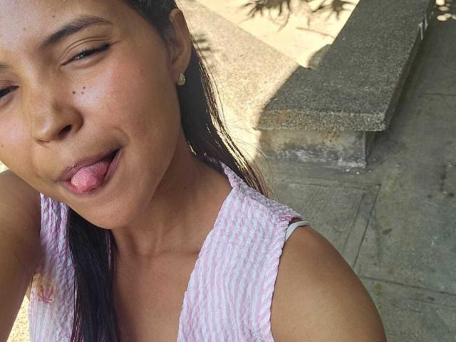 Close-up of MelaniaVelasquez with medium brown skin, sticking out her tongue slightly, wearing a white, textured tank top. Background includes concrete and shadowed areas. She has a playful expression.