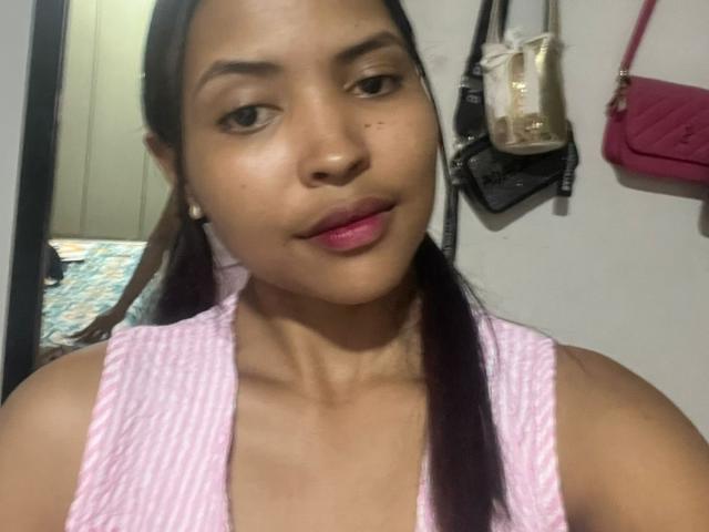 Foto van Melania Velasquez met lang zwart haar in vlechtjes, die een roze en witte gestreepte tanktop draagt, roze lipsticks en kleine oorbellen. Koffers hangen aan een witte muur op de achtergrond. Verdomd aantrekkelijk..