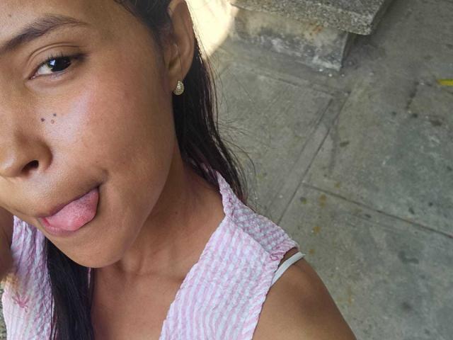 Primo piano di Melania Velasquez con la pelle olivastra, i capelli neri e piccoli orecchini, che mostra la lingua. Indossa una camicetta bianca con un motivo, con un muro di pietra strutturato sullo sfondo. Molto attraente..