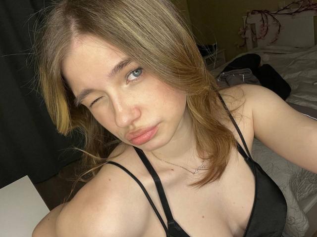 Close-up of JennyOsmun with light brown hair, winking, wearing a black spaghetti strap top, exposing cleavage. She has fair skin and a slight smirk, with a subtle necklace. Bedroom setting.