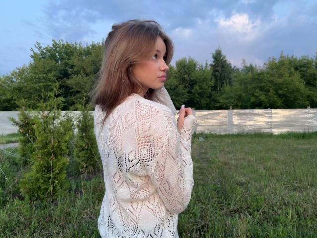 JennyOsmun se tient dans un champ herbeux, vêtue d'un pull blanc orné de motifs en dentelle, et regarde par-dessus son épaule avec un léger faire la moue.
elle a les cheveux bruns et dégage une aura séduisante. On peut voir des arbres et le ciel en arrière-plan..